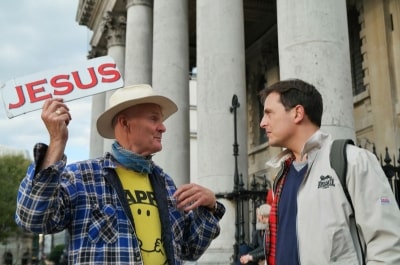 Photo of man holding sign of Jesus on street and in discussion with another man.